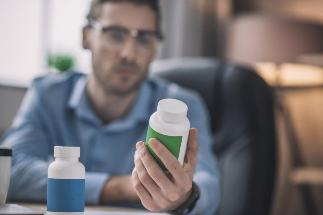 Man in a blue shirt and glasses examining the back of a supplement bottle as his desk.
