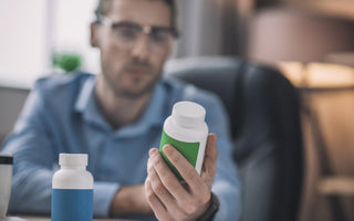 Man in a blue shirt and glasses examining the back of a supplement bottle as his desk.