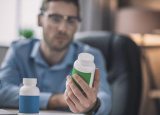 Man in a blue shirt and glasses examining the back of a supplement bottle as his desk.