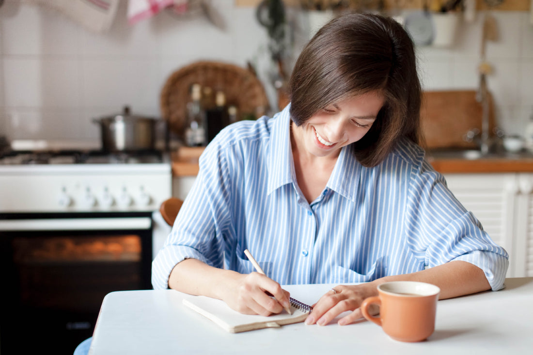 Woman in a blue and white striped blouse sitting at a kitchen table writing in a notebook.