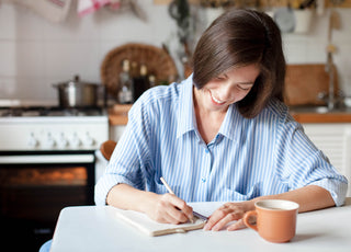 Woman in a blue and white striped blouse sitting at a kitchen table writing in a notebook.