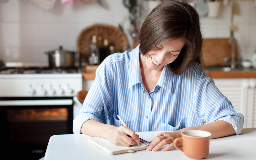 Woman in a blue and white striped blouse sitting at a kitchen table writing in a notebook.