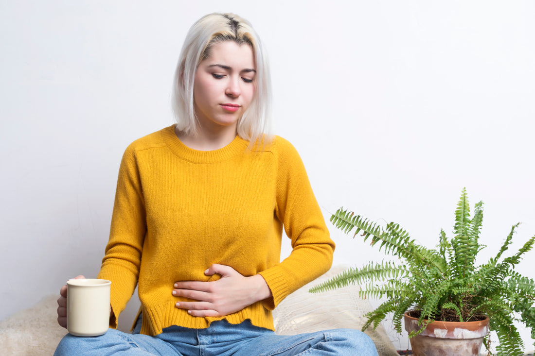Woman with short blond hair wearing jeans and a yellow sweater sitting cross-legged on the floor holding a coffee mug in one hand with her other hand placed over her stomach.