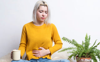 Woman with short blond hair wearing jeans and a yellow sweater sitting cross-legged on the floor holding a coffee mug in one hand with her other hand placed over her stomach.