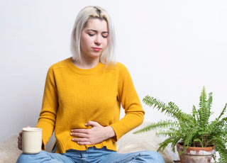 Woman with short blond hair wearing jeans and a yellow sweater sitting cross-legged on the floor holding a coffee mug in one hand with her other hand placed over her stomach.