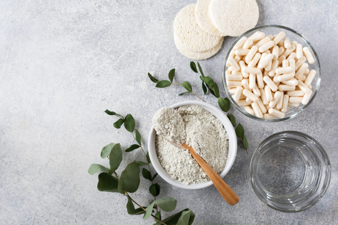 Flatlay of a bowl of white powder with a wooden spoon laying across it, a glass bowl full of white supplements, and a glass of water on a gray stone countertop. 