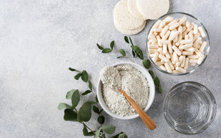 Flatlay of a bowl of white powder with a wooden spoon laying across it, a glass bowl full of white supplements, and a glass of water on a gray stone countertop. 