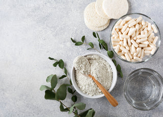 Flatlay of a bowl of white powder with a wooden spoon laying across it, a glass bowl full of white supplements, and a glass of water on a gray stone countertop. 