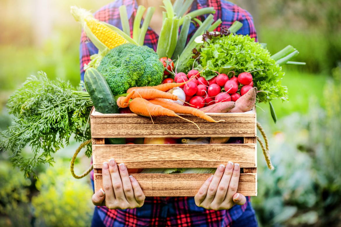 Person in a red and blue plaid shirt holding a wooden crate full of fresh vegetables.