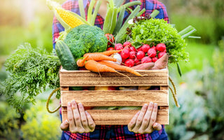 Person in a red and blue plaid shirt holding a wooden crate full of fresh vegetables.