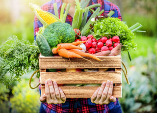 Person in a red and blue plaid shirt holding a wooden crate full of fresh vegetables.