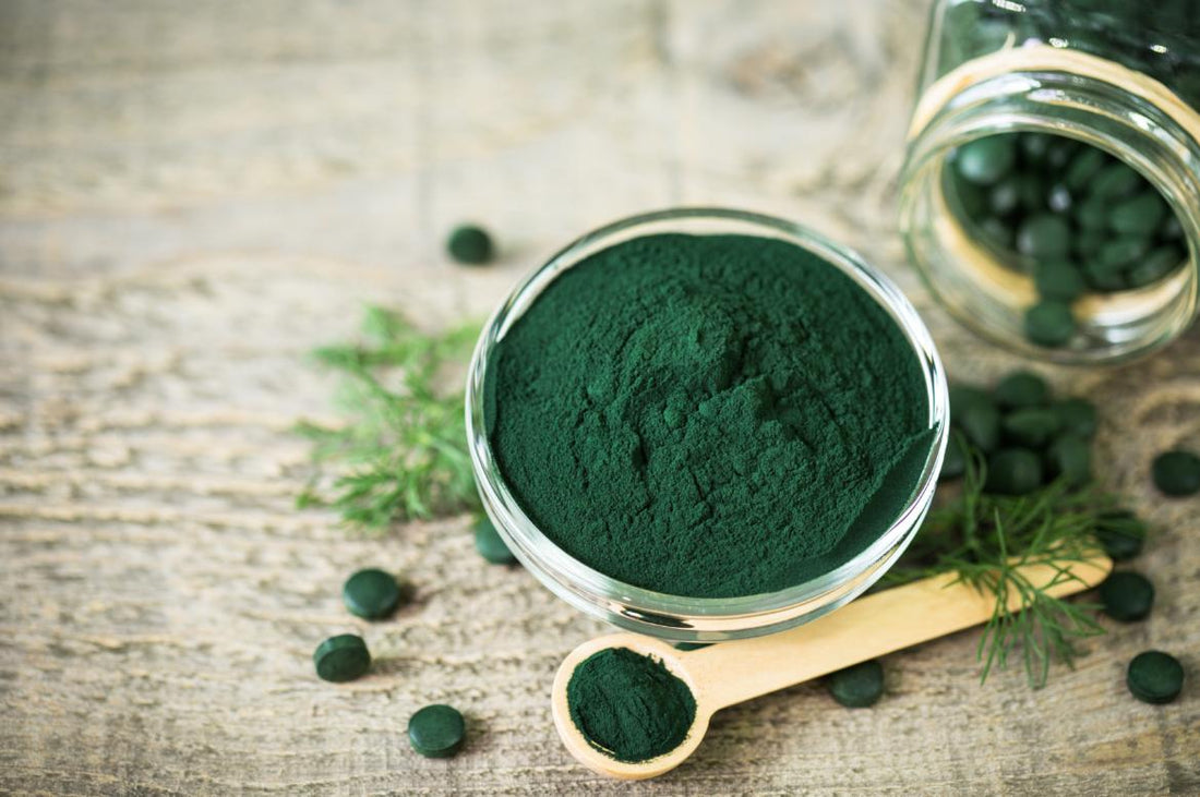 A clear bowl full of green algae powder is sitting on a wooden tabletop next to a measuring spoon full of the same powder and few green tablets.