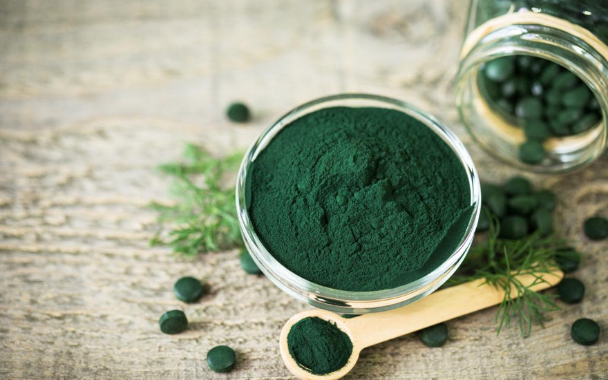 A clear bowl full of green algae powder is sitting on a wooden tabletop next to a measuring spoon full of the same powder and few green tablets.