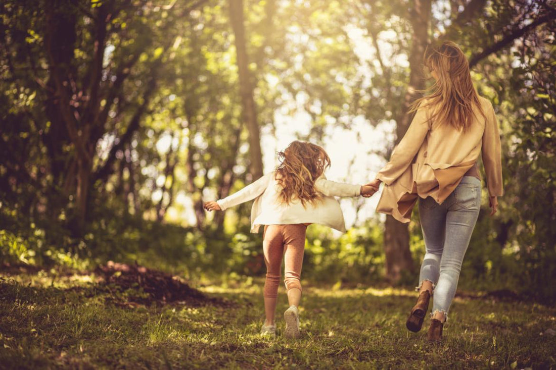 Mother with her young daughter holding hands and walking through a lightly forested area.