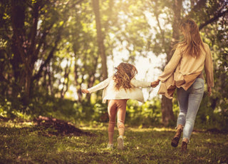 Mother with her young daughter holding hands and walking through a lightly forested area.