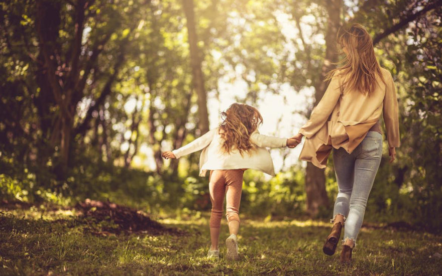 Mother with her young daughter holding hands and walking through a lightly forested area.