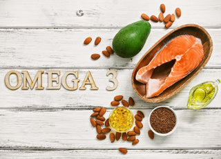 Wooden letters spelling "OMEGA 3" laying on a wooden table surrounded by fatty foods like salmon and avocado.