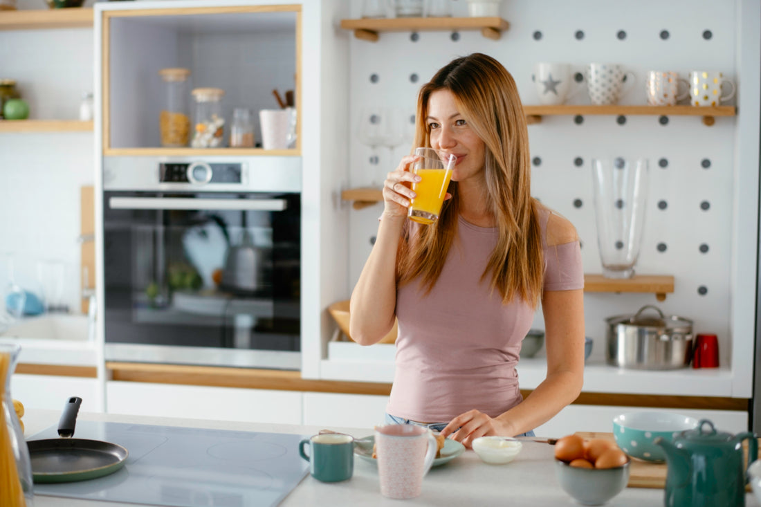 Woman in a pink shirt standing at a kitchen counter sipping from a glass of orange juice.