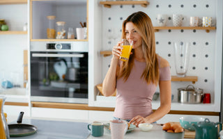 Woman in a pink shirt standing at a kitchen counter sipping from a glass of orange juice.