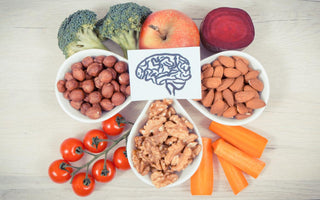 A piece of paper with a drawing of a brain on it sitting on top of an array of healthy foods including apples, broccoli, bowls of nuts, tomoatos, and peppers.