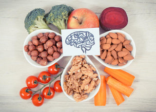 A piece of paper with a drawing of a brain on it sitting on top of an array of healthy foods including apples, broccoli, bowls of nuts, tomoatos, and peppers.