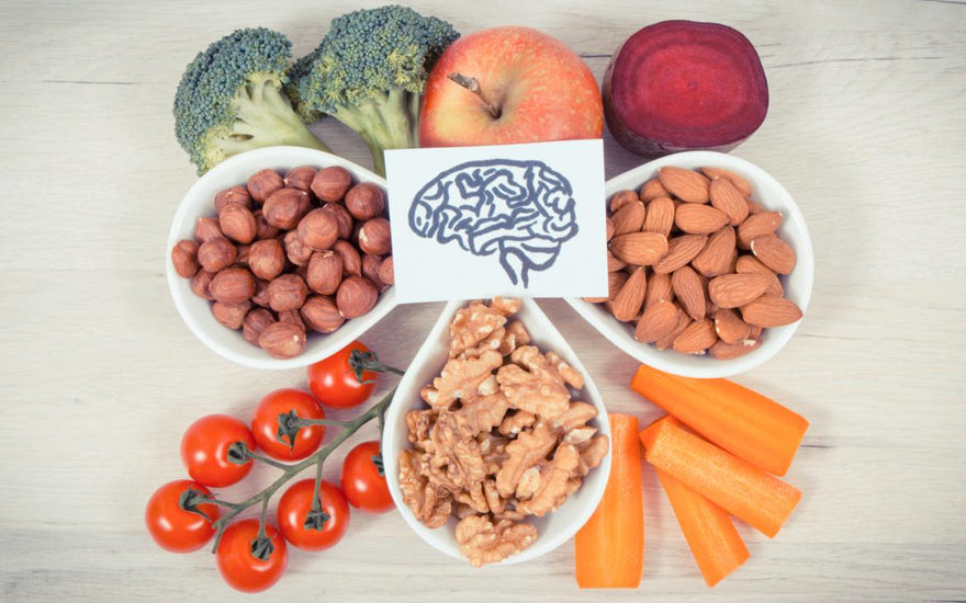 A piece of paper with a drawing of a brain on it sitting on top of an array of healthy foods including apples, broccoli, bowls of nuts, tomoatos, and peppers.