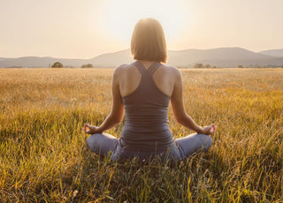 Woman sitting cross-legged with her back to the camera in a field of grass as the sun sets.