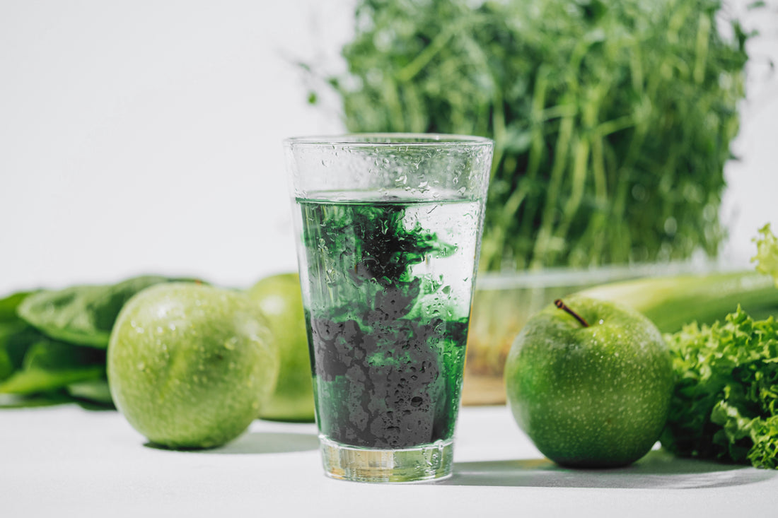 Glass of water with chlorophyll drops dissolving in it sitting on a counter surrounded by green produce such as apples and celery. 