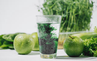 Glass of water with chlorophyll drops dissolving in it sitting on a counter surrounded by green produce such as apples and celery. 