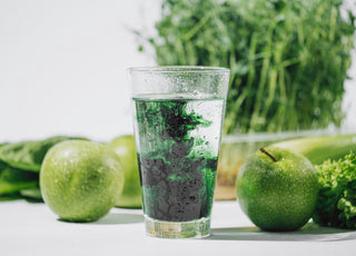 Glass of water with chlorophyll drops dissolving in it sitting on a counter surrounded by green produce such as apples and celery. 