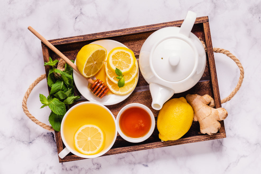 Wooden tray filled with a white tea kettle and a cup of tea as well as fresh honey, ginger, and lemon. 