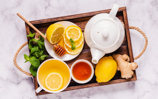 Wooden tray filled with a white tea kettle and a cup of tea as well as fresh honey, ginger, and lemon. 