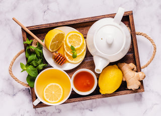 Wooden tray filled with a white tea kettle and a cup of tea as well as fresh honey, ginger, and lemon. 