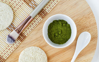 Flat-lay of beauty products on a wooden tray, including a makeup brush, pumice stone, and white bowl filled with blue-green algae powder.