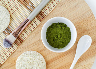 Flat-lay of beauty products on a wooden tray, including a makeup brush, pumice stone, and white bowl filled with blue-green algae powder.