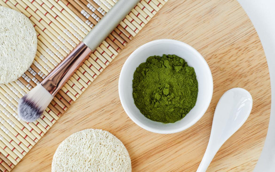 Flat-lay of beauty products on a wooden tray, including a makeup brush, pumice stone, and white bowl filled with blue-green algae powder.