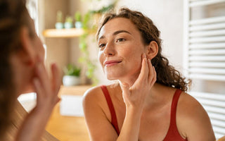 Young woman with her hair pulled back looking in the mirror with her fingers resting on her jawline.