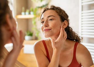 Young woman with her hair pulled back looking in the mirror with her fingers resting on her jawline.