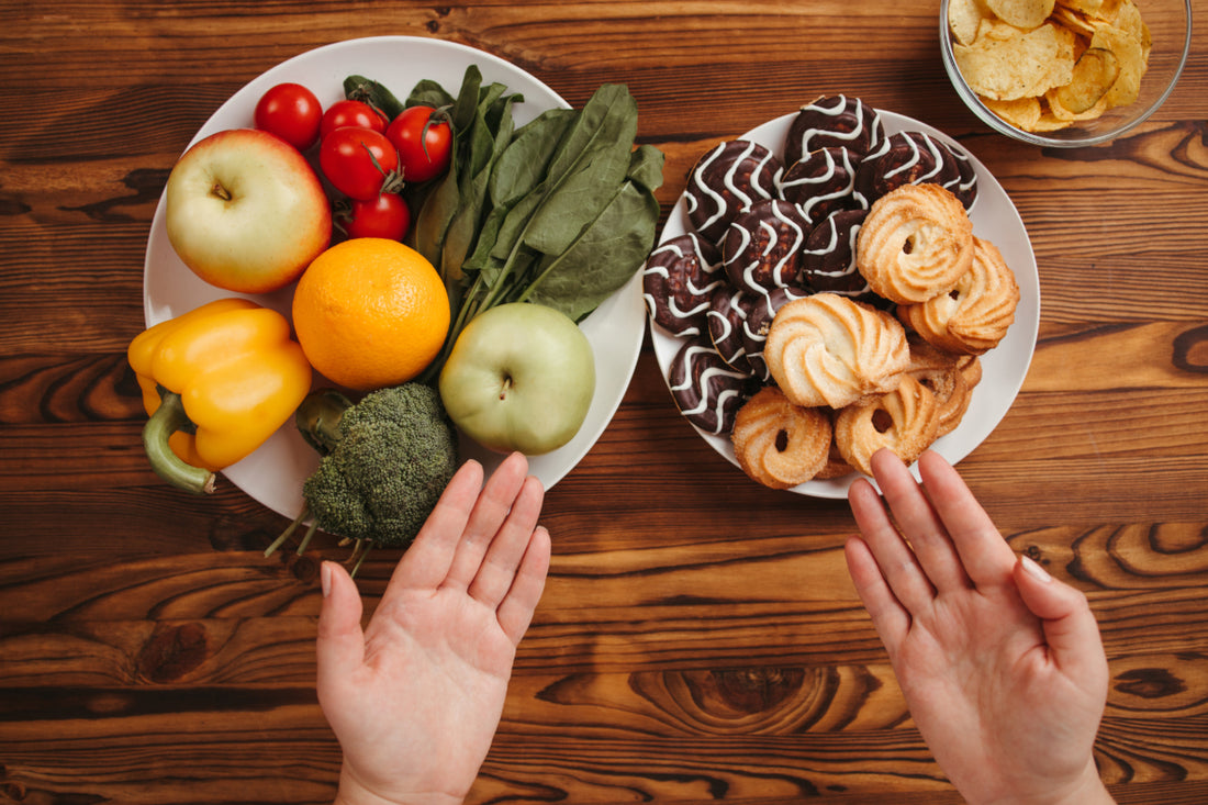Two hands gesturing toward two different plates on a wooden tabletop. One plate is full of fresh fruits and vegetables while the other has two different types of cookies on it.