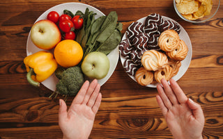 Two hands gesturing toward two different plates on a wooden tabletop. One plate is full of fresh fruits and vegetables while the other has two different types of cookies on it.