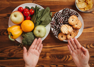 Two hands gesturing toward two different plates on a wooden tabletop. One plate is full of fresh fruits and vegetables while the other has two different types of cookies on it.