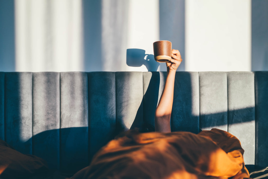 Person laying in bed with an orange comforter pulled over their head while they hold a brown coffee mug up in the air.