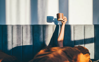 Person laying in bed with an orange comforter pulled over their head while they hold a brown coffee mug up in the air.