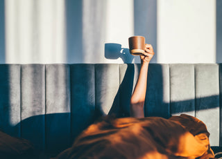 Person laying in bed with an orange comforter pulled over their head while they hold a brown coffee mug up in the air.