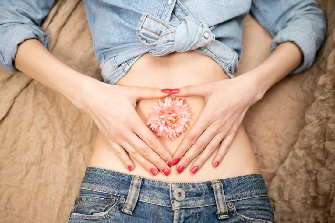 Woman laying on her back with her stomach exposed, a pink flower placed over her bellybutton, and her hands placed gently around the flower.