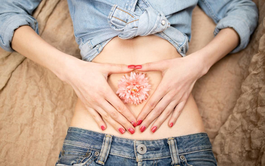 Woman laying on her back with her stomach exposed, a pink flower placed over her bellybutton, and her hands placed gently around the flower.