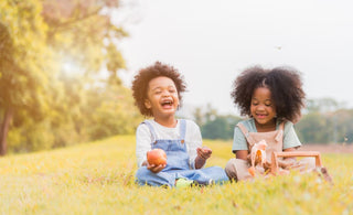 Two young children sitting in the grass while snacking on fruit. 