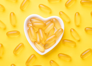 A white heart-shaped bowel filled with fish oil supplements sitting on a yellow background. 