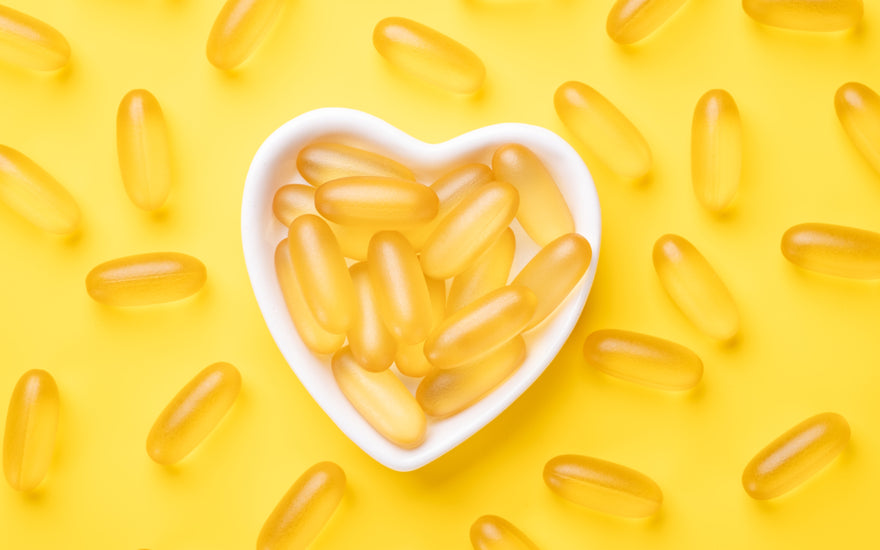 A white heart-shaped bowel filled with fish oil supplements sitting on a yellow background. 