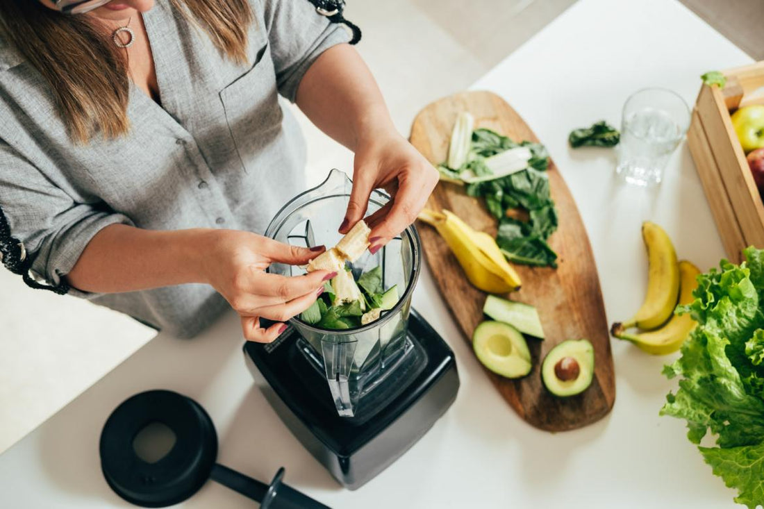 Top-down view of a woman breaking a banana in half so she can add it to the blender that is already partly full of smoothie ingredients.